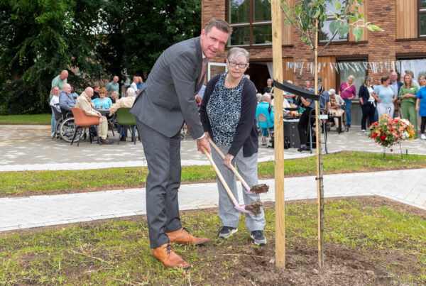 Wethouder en client planten appelboom. Ze hebben allebei een roze schepje in de hand.