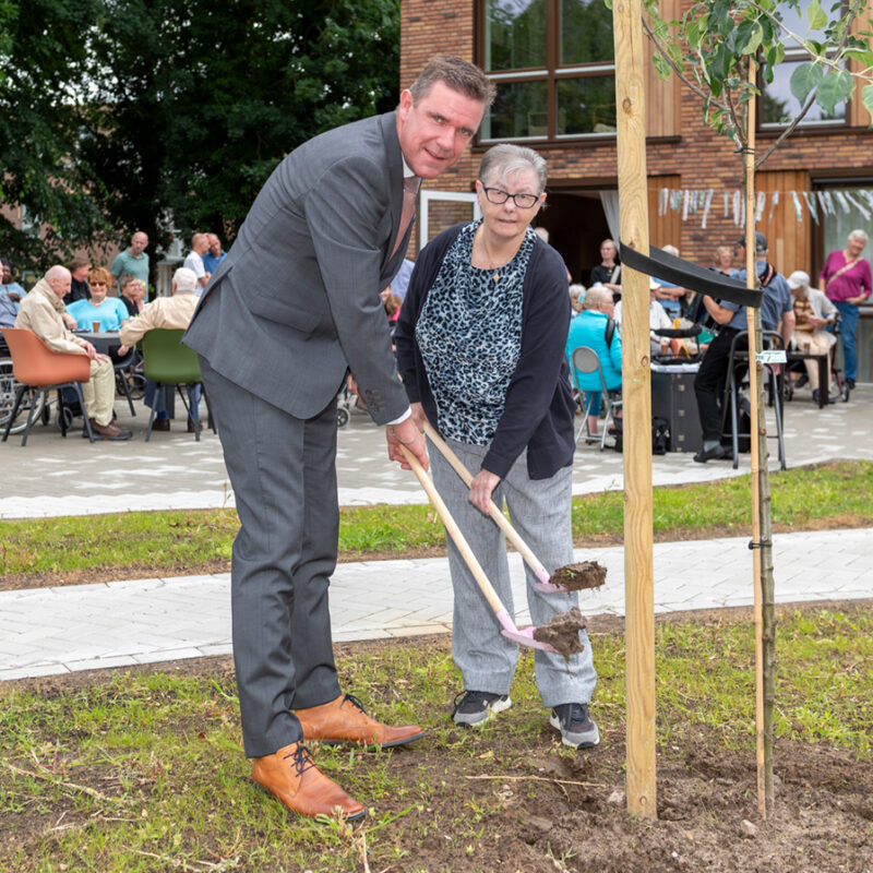 Wethouder en client planten appelboom. Ze hebben allebei een roze schepje in de hand.